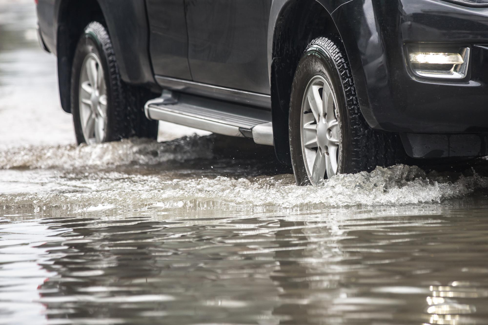 SUV preta passando por uma rua alagada, com água cobrindo parte dos pneus durante uma chuva ou enchente urbana.